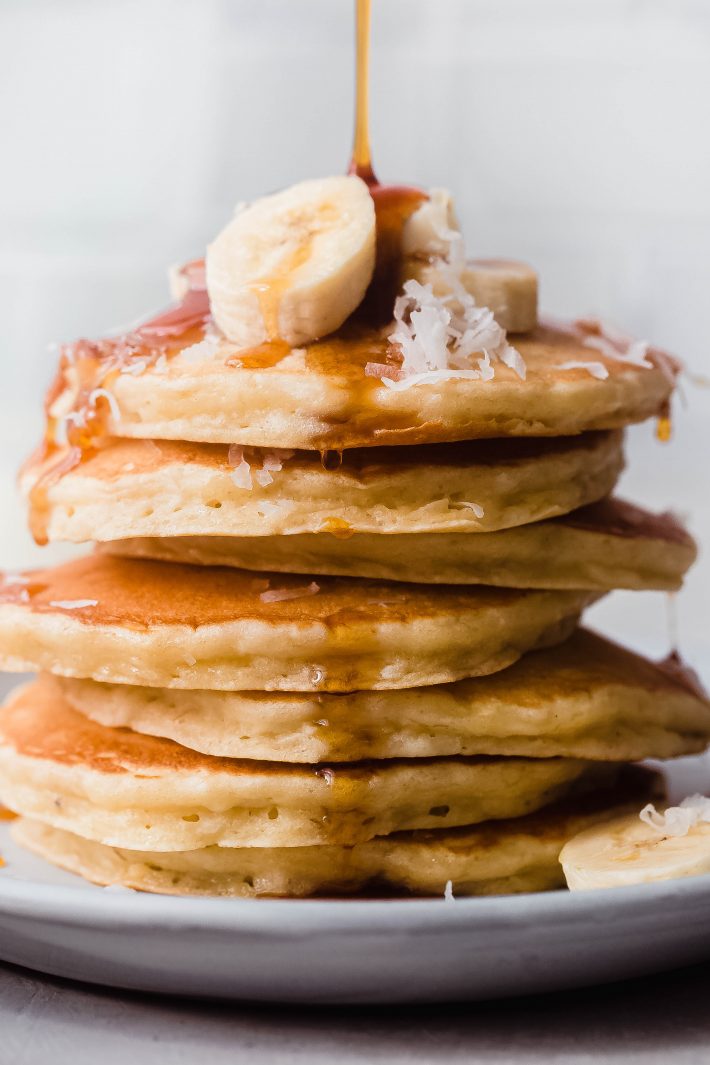 syrup pouring over pancakes with banana slices and shredded coconut