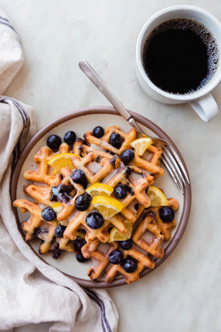 plateful of waffles with lemon wedges and blueberries on white marble with fork and coffee