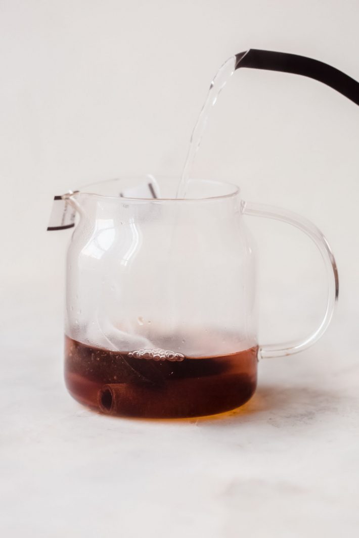 hot water being poured into glass tea pot with tea bags and cinnamon stick
