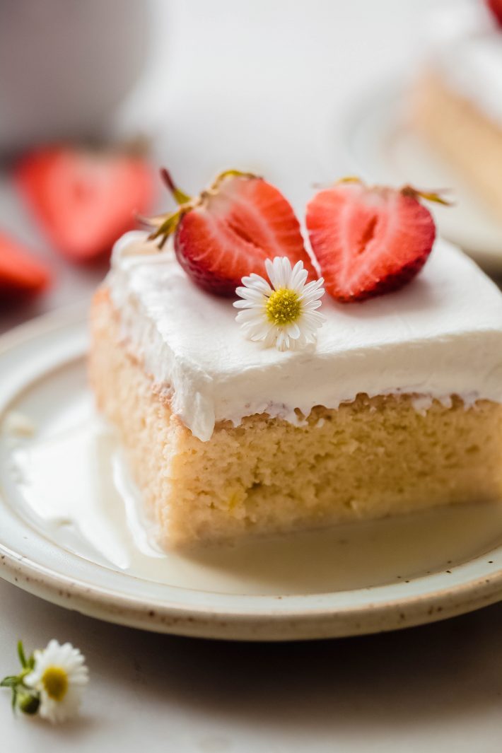 slice of tres leches cake on plate with milk syrup, whipped cream, strawberries, and a small flower